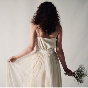 May include: A white wedding dress with a lace belt and a flowing skirt. The dress is being worn by a person with long brown hair.