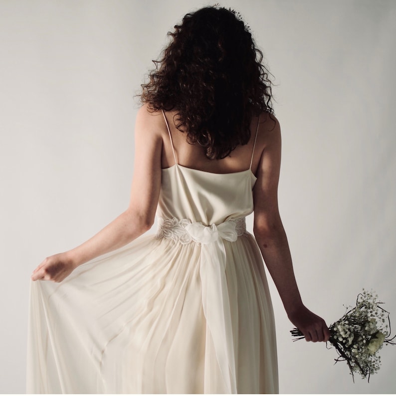 May include: A white wedding dress with a lace belt and a flowing skirt. The dress is being worn by a person with long brown hair.