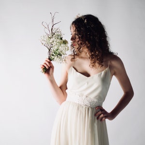 May include: A woman in a white, sleeveless wedding dress with a delicate lace belt holds a bouquet of small white flowers.