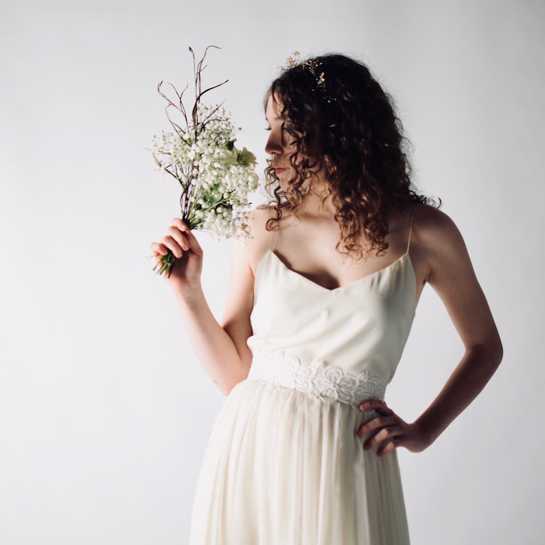 May include: A woman in a white, sleeveless wedding dress with a delicate lace belt holds a bouquet of small white flowers.