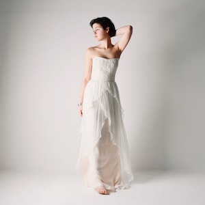Op de afbeelding: Een witte strapless bruidsjurk met een gelaagde rok en een gerimpelde zoom. De jurk is gemaakt van een transparante stof en heeft een aansluitend lijfje.