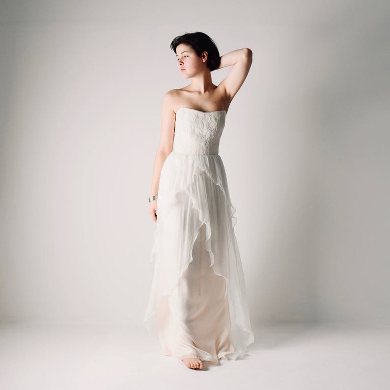 Op de afbeelding: Een witte strapless bruidsjurk met een gelaagde rok en een gerimpelde zoom. De jurk is gemaakt van een transparante stof en heeft een aansluitend lijfje.