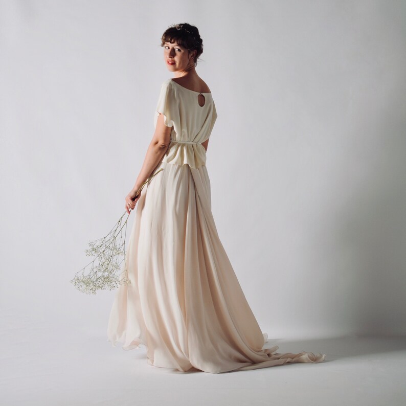 May include: A cream-colored wedding dress with a flowing skirt and short sleeves. The dress features a keyhole back and a cinched waist. The model is holding a bouquet of white flowers. The dress is made of a soft, lightweight fabric.