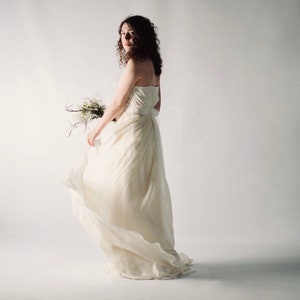 May include: A woman in a white wedding dress with a flowing skirt, holding a bouquet of white flowers. She is standing in front of a white background.