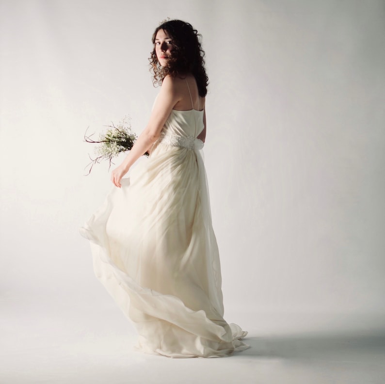 May include: A woman in a white wedding dress with a flowing skirt, holding a bouquet of white flowers. She is standing in front of a white background.