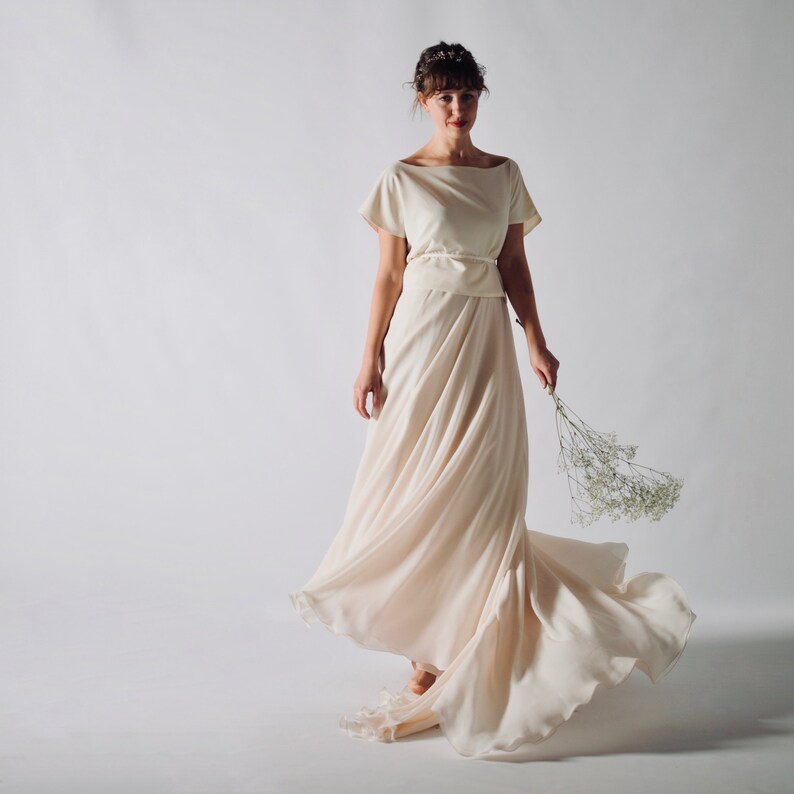 May include: A cream-colored wedding dress with a boat neckline and short sleeves. The dress features a flowing skirt and a matching belt. The model is holding a small bouquet of white flowers. The dress is made of a soft, flowing fabric.