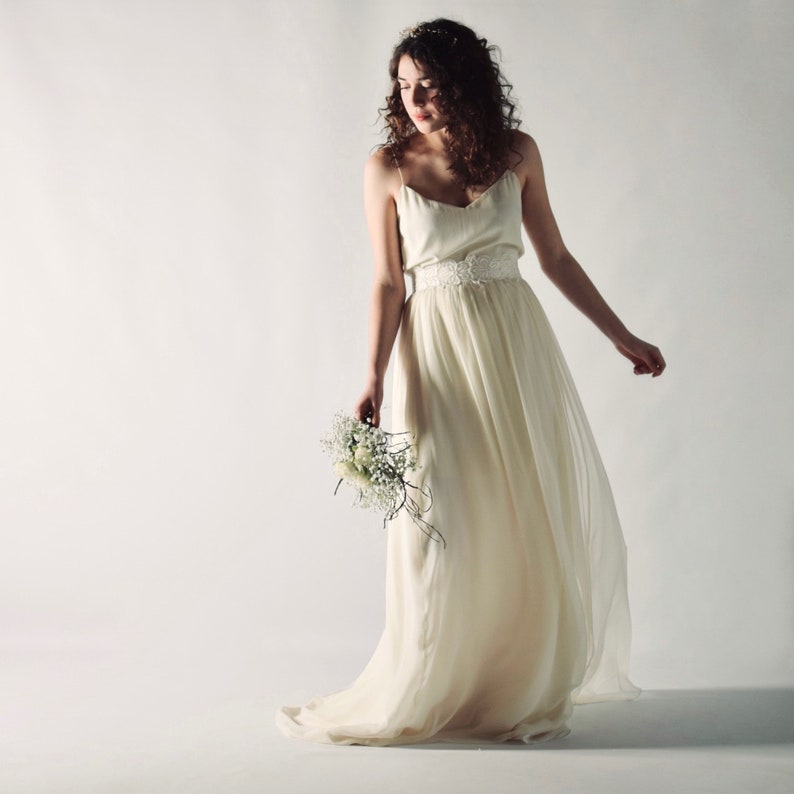 May include: A woman in a white, flowing wedding dress with a lace belt and a bouquet of white flowers.