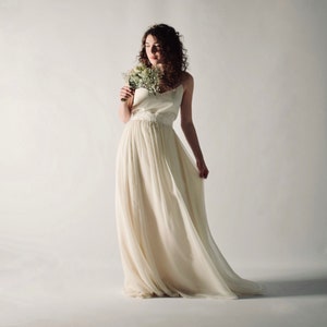 May include: A woman in a white wedding dress with a lace bodice and a flowing skirt. She is holding a bouquet of white flowers and looking down at them.