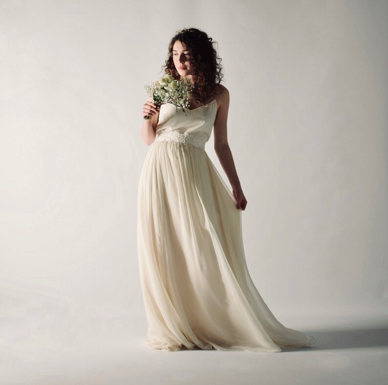 May include: A woman in a white wedding dress with a lace bodice and a flowing skirt. She is holding a bouquet of white flowers and looking down at them.