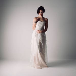 Op de afbeelding: Een vrouw in een witte strapless bruidsjurk met een gerimpelde rok en een delicate kanten bustier. Ze staat in een studio met een witte achtergrond.