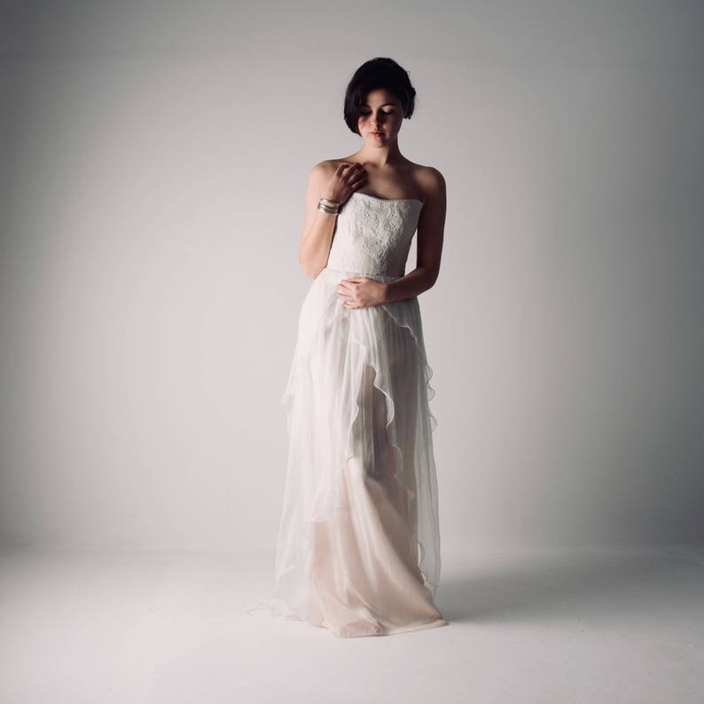 Op de afbeelding: Een vrouw in een witte strapless bruidsjurk met een gerimpelde rok en een delicate kanten bustier. Ze staat in een studio met een witte achtergrond.