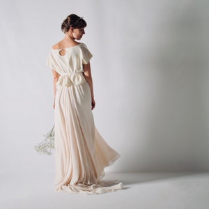 May include: A cream-colored wedding dress with a short-sleeved top and a flowing skirt. The top features a keyhole back and a cinched waist. The dress is paired with a delicate floral headpiece and a small bouquet of white flowers.