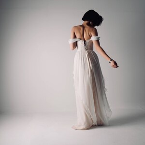 Op de afbeelding: Een vrouw draagt een witte off-shoulder jurk met een veter aan de achterkant. De jurk heeft een zwierige rok en is gemaakt van een lichtgewicht stof.