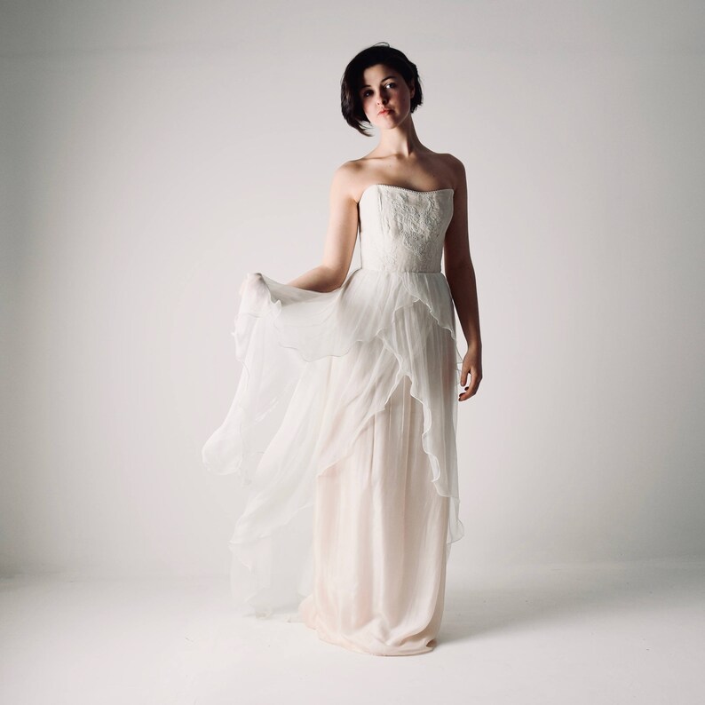 Op de afbeelding: Een vrouw die een witte strapless bruidsjurk draagt met een zwierige rok en lagen tule. De jurk heeft een aansluitend lijfje en een eenvoudig ontwerp.