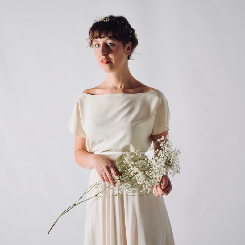 May include: A cream-colored dress with a boat neckline and short sleeves. The dress is paired with a delicate floral crown and a bouquet of white flowers. The overall aesthetic is soft and elegant, suitable for a wedding or special occasion.