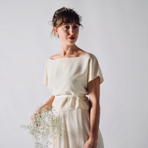 May include: Cream-colored, two-piece bridal outfit. The top has a boat neckline and short sleeves. The skirt is long and flowing. A thin belt cinches the waist. The outfit is paired with a delicate floral hairpiece and a bouquet of white flowers.