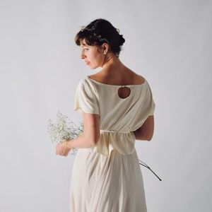 May include: A cream-colored dress with a keyhole back and a flowing skirt. The dress features short sleeves and a fitted waist. The person is holding a bouquet of white flowers and wearing a delicate floral headpiece. The dress is a soft, neutral color.