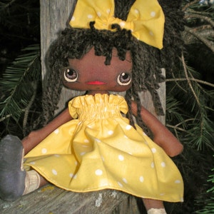 Puede incluir: Una muñeca de trapo hecha a mano con pelo de lana negro, un vestido amarillo con lunares blancos y un lazo amarillo con lunares blancos.