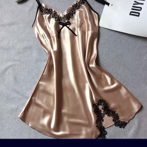 May include: A champagne-coloured satin slip dress with black lace trim. The dress features thin straps, a V-neckline, and a small bow detail. The lace trim is along the neckline and hem.