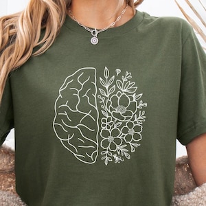 Op de afbeelding: Olijfgroen T-shirt met een wit grafisch ontwerp van een hersenhelft en bloemen. Het ontwerp is verdeeld, met de hersenen aan de ene kant en bloemen aan de andere kant. Het woord "Military" staat op een blauwe banner. Het T-shirt is van zacht materiaal.