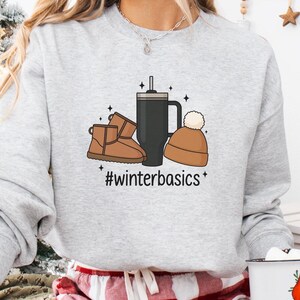 May include: A light gray sweatshirt with a graphic of winter boots, a travel mug, and a beanie. The text "#winterbasics" is below the graphic. The person is wearing red and white plaid pajama pants and holding a white mug with a Santa Claus graphic.