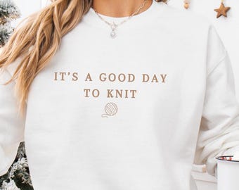 May include: White long-sleeved sweatshirt with the text "IT'S A GOOD DAY TO KNIT" and a yarn ball graphic. The image also shows a white mug with a Santa Claus illustration. The word "White" is displayed in a teal banner.