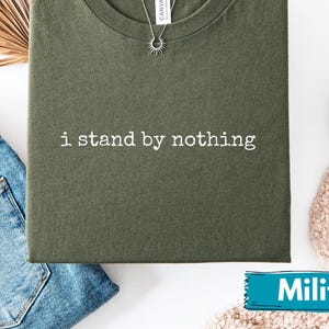 Może przedstawiać: Zielona koszulka w kolorze oliwkowym z białym napisem "i stand by nothing". Koszulka jest złożona i umieszczona z niebieskimi dżinsami, białymi trampkami i srebrnym naszyjnikiem. Na zdjęciu znajduje się również suszony liść palmy.