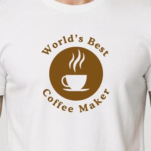 Può includere: T-shirt bianco con grafica circolare marrone con la scritta "World's Best Coffee Maker" attorno a una tazza di caffè fumante. La maglietta è a girocollo e ha maniche corte.