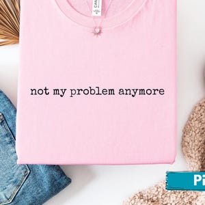 Könnte beinhalten: Ein hellrosa T-Shirt mit dem schwarzen Aufdruck "not my problem anymore". Das Shirt liegt gefaltet auf einer weißen Oberfläche, neben einer blauen Jeans und weißen Schuhen. Eine Halskette ist sichtbar.