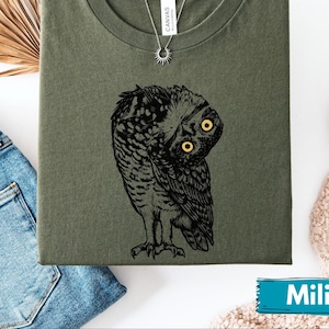 May include: An olive green t-shirt with a black and white owl illustration and yellow eyes. The shirt is folded, with a silver necklace and sun pendant. The word "Military" is on a blue banner. The shirt is a crew neck.