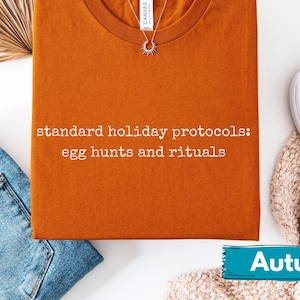 Könnte beinhalten: Ein orangefarbenes T-Shirt mit dem Text "standard holiday protocols: egg hunts and rituals". Das Shirt wird mit blauen Jeans, weißen Turnschuhen und einer Halskette kombiniert. Ein Banner unten zeigt "Autumn".