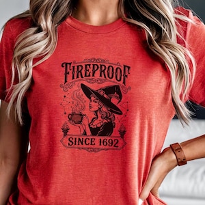 Puede incluir: Camiseta roja con un diseño negro de una bruja sosteniendo una taza, con el texto "FIREPROOF SINCE 1692". La bruja lleva un sombrero. La camiseta de manga corta es de color rojo vivo.