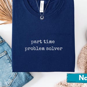 Può includere: T-shirt blu navy con la scritta bianca "part time problem solver". Una collana d'argento con un ciondolo a forma di sole è appoggiata sulla maglietta. La maglietta è piegata accanto a jeans blu e scarpe bianche.