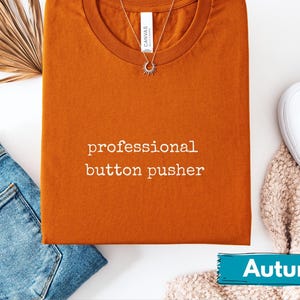 May include: An orange t-shirt with the text "professional button pusher" in white, paired with blue jeans and white sneakers. A gold necklace with a sun pendant is on the shirt. The word "Autumn" is on a teal banner.