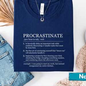 May include: Navy blue t-shirt with the definition of "Procrastinate" printed in white. The definition includes the pronunciation, the word's part of speech, and three numbered points. A silver necklace with a sunburst pendant is visible.