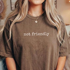 May include: Espresso-colored t-shirt with the text "not friendly" in white. The shirt is made to order and is a relaxed fit. The model is holding a mug.