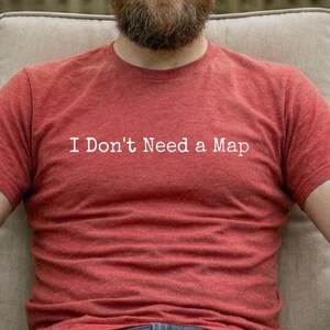 May include: A red t-shirt with the text "I Don't Need a Map" in white. The person is wearing blue jeans and sunglasses while sitting in a chair outdoors. The background includes trees and a wooden fence.