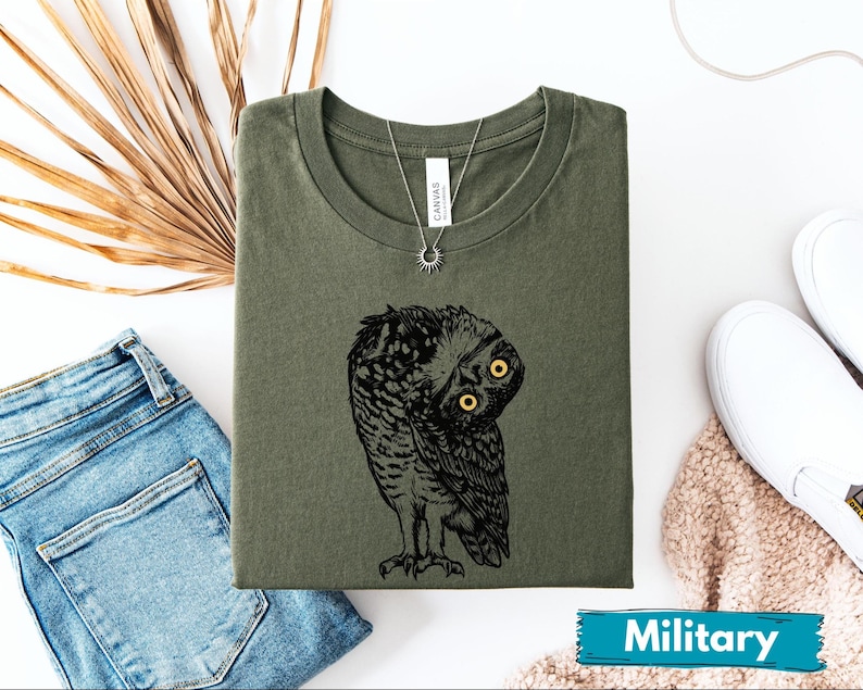 May include: An olive green t-shirt with a black and white owl illustration and yellow eyes. The shirt is folded, with a silver necklace and sun pendant. The word "Military" is on a blue banner. The shirt is a crew neck.