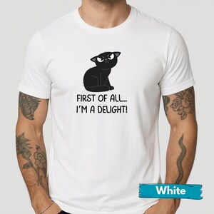 May include: White t-shirt featuring a black cartoon cat with an angry expression. The text "FIRST OF ALL... I'M A DELIGHT!" is printed below the cat. The shirt has short sleeves and a crew neck.