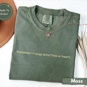 May include: A moss green short-sleeve t-shirt with a crew neckline and a Comfort Colors label. The shirt features yellow printed text that reads: if (doorbell == rung): print("Trick or Treat!"). A thin gold-toned necklace rests on the fabric.