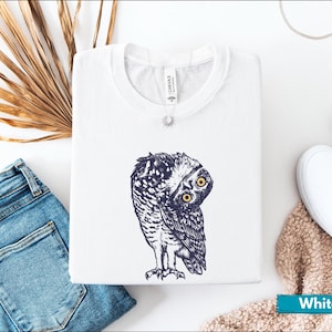 May include: White t-shirt with a navy blue and white owl illustration, featuring yellow eyes. The folded shirt is on a white surface, alongside blue jeans and white trainers. The shirt has a 'Canvas' tag.