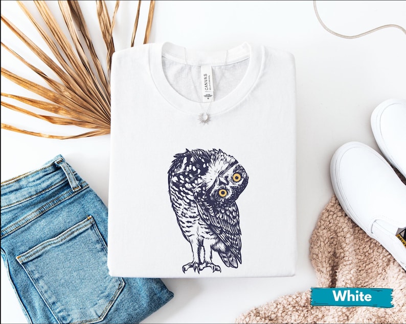 May include: White t-shirt with a navy blue and white owl illustration, featuring yellow eyes. The folded shirt is on a white surface, alongside blue jeans and white trainers. The shirt has a 'Canvas' tag.