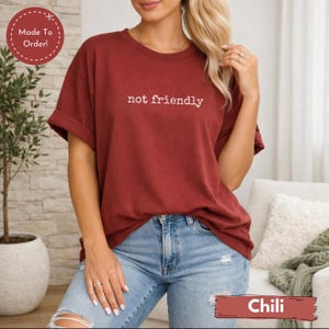 May include: A maroon-colored t-shirt with the words "not friendly" printed in white. The shirt is paired with blue jeans. The bottom right corner of the image has the word "Chili" in a red box. The shirt is made to order.