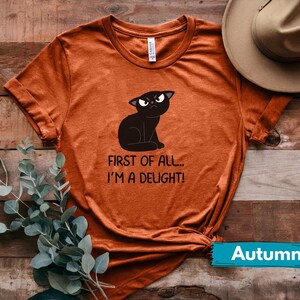 May include: Burnt orange t-shirt featuring a black cat illustration with an angry expression. The text reads "FIRST OF ALL... I'M A DELIGHT!". A hat and greenery are visible in the background. The word "Autumn" is on a teal banner.