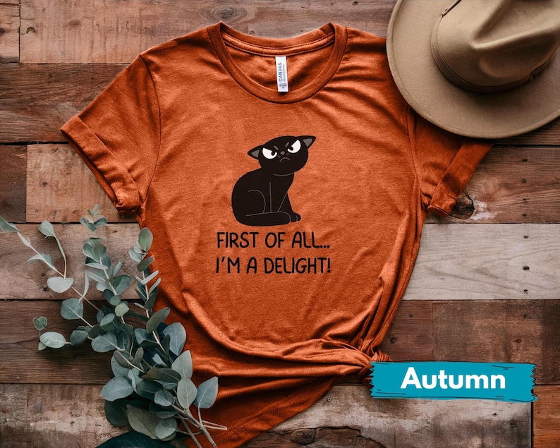 May include: Burnt orange t-shirt featuring a black cat illustration with an angry expression. The text reads "FIRST OF ALL... I'M A DELIGHT!". A hat and greenery are visible in the background. The word "Autumn" is on a teal banner.
