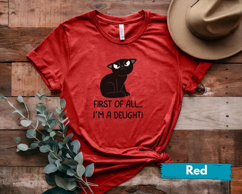 May include: Red t-shirt featuring a black cat graphic with an angry expression. The text on the shirt reads "FIRST OF ALL... I'M A DELIGHT!". The shirt is made of a soft material and is a casual wear item.