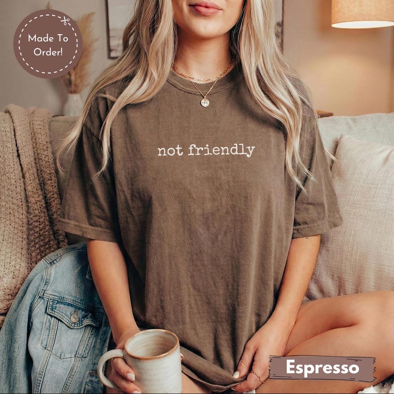 May include: Espresso-colored t-shirt with the text "not friendly" in white. The shirt is made to order and is a relaxed fit. The model is holding a mug.