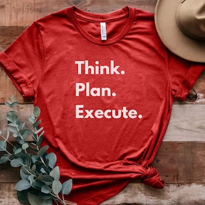 Puede incluir: Una camiseta roja con las palabras "Think. Plan. Execute." en letras blancas. La camiseta está anudada en la parte inferior y descansa sobre una superficie de madera con vegetación y un sombrero beige.