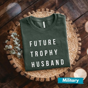 May include: A dark green t-shirt with the words "FUTURE TROPHY HUSBAND" printed in white. The shirt is displayed on a woven, circular placemat with a sprig of greenery, set against a dark wood background.