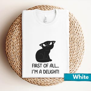 May include: White t-shirt featuring a cartoon black cat with an angry expression. The text "FIRST OF ALL... I'M A DELIGHT!" is printed below the cat. A teal banner at the bottom reads "White".